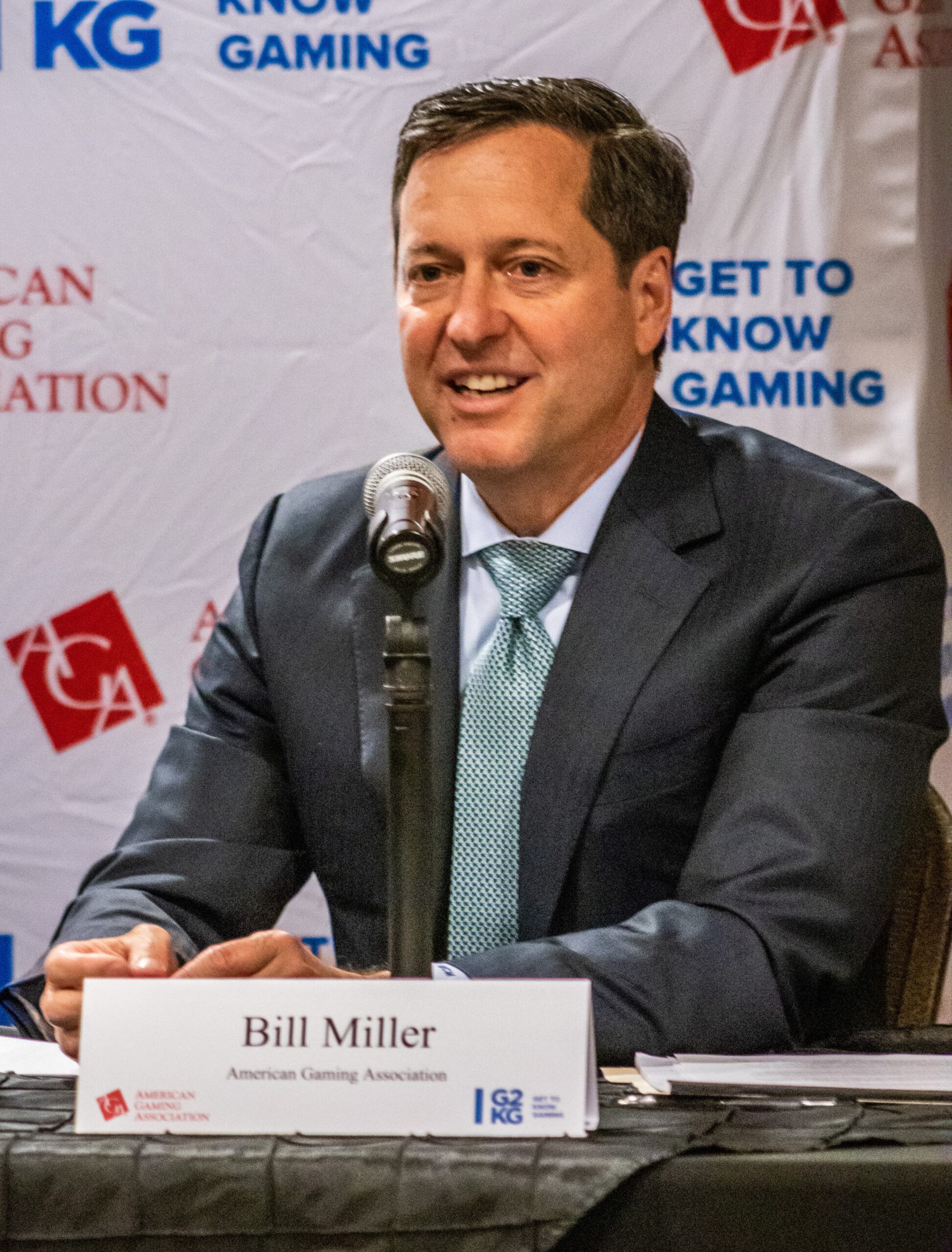 A man in a suit speaks into a microphone at a conference or event. A nameplate reads "Bill Miller" with the American Gaming Association logo beside it. The backdrop displays various logos including "GET TO KNOW GAMING" and "AGA.