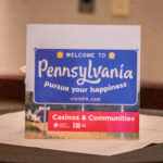 A promotional sign sits on a table, featuring a photo of a Pennsylvania welcome sign with the text "WELCOME TO Pennsylvania, pursue your happiness." The bottom section reads "Casinos & Communities" with logos.