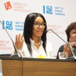 Two women sit at a conference table with microphones. The woman on the left gestures with her hands, wearing a white blouse and pearls. The backdrop features logos for the American Gaming Association and "Get to Know Gaming.