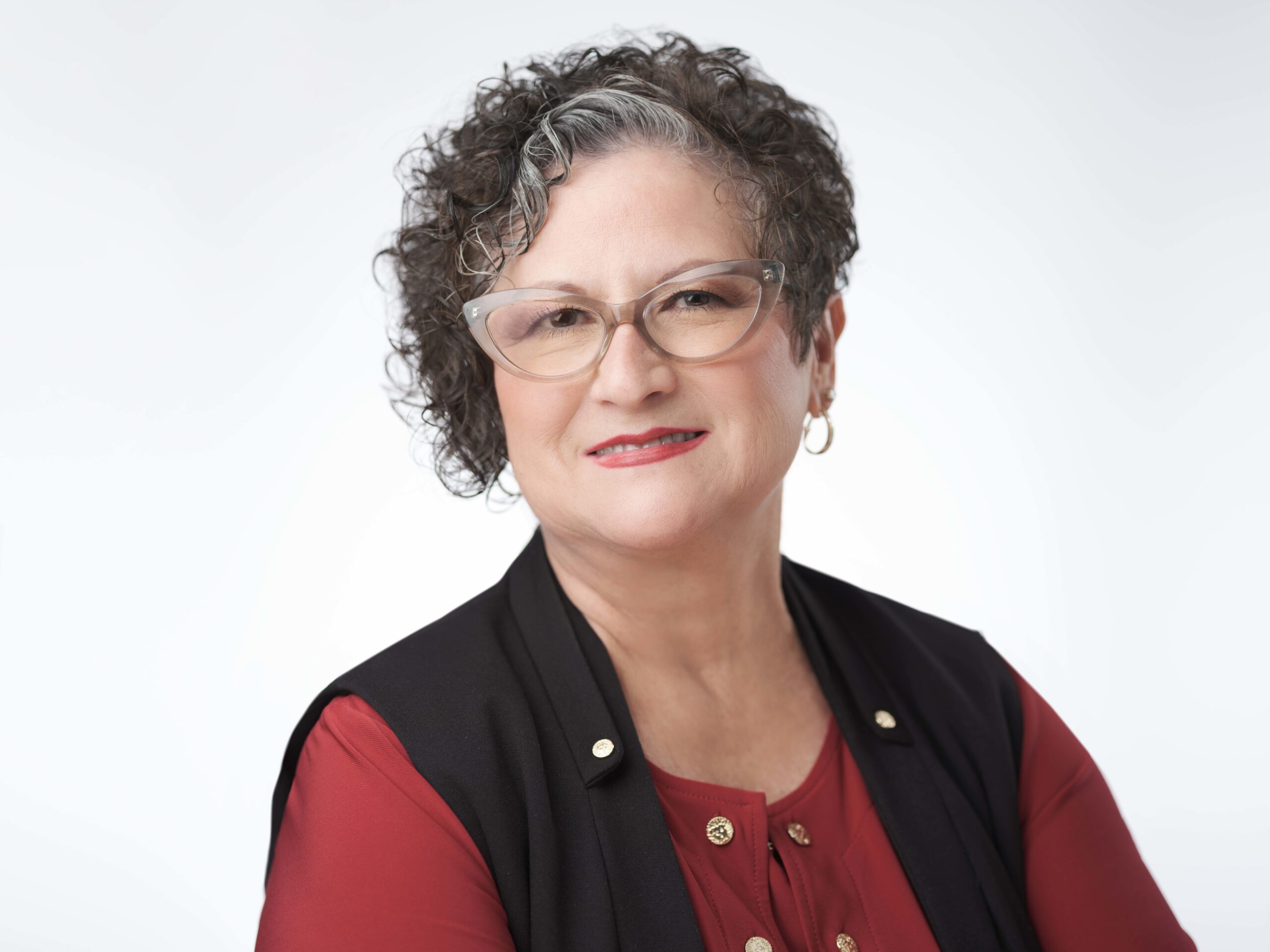 A woman with curly hair, wearing glasses, a red blouse, and a black vest smiles at the camera. She has gold earrings and two gold buttons on her blouse. The background is plain white.