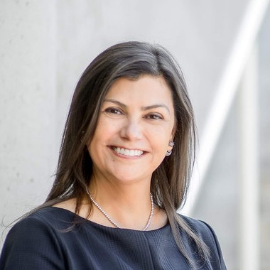 A woman with long dark hair is smiling while standing against a light gray background. She is wearing a black top and a silver necklace.