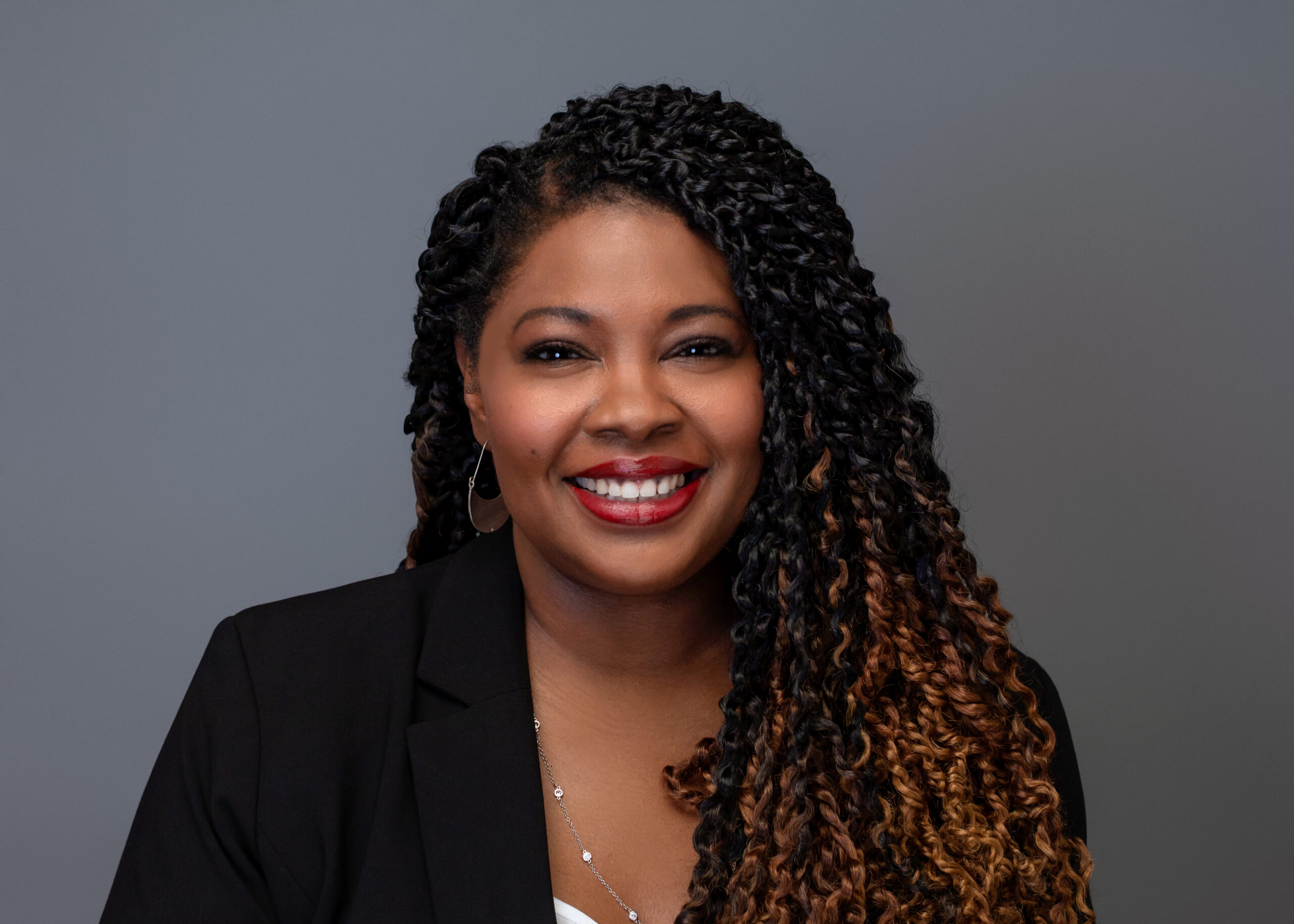 A person with long braided hair, smiling, dressed in a black blazer and a white top, posed against a gray background.