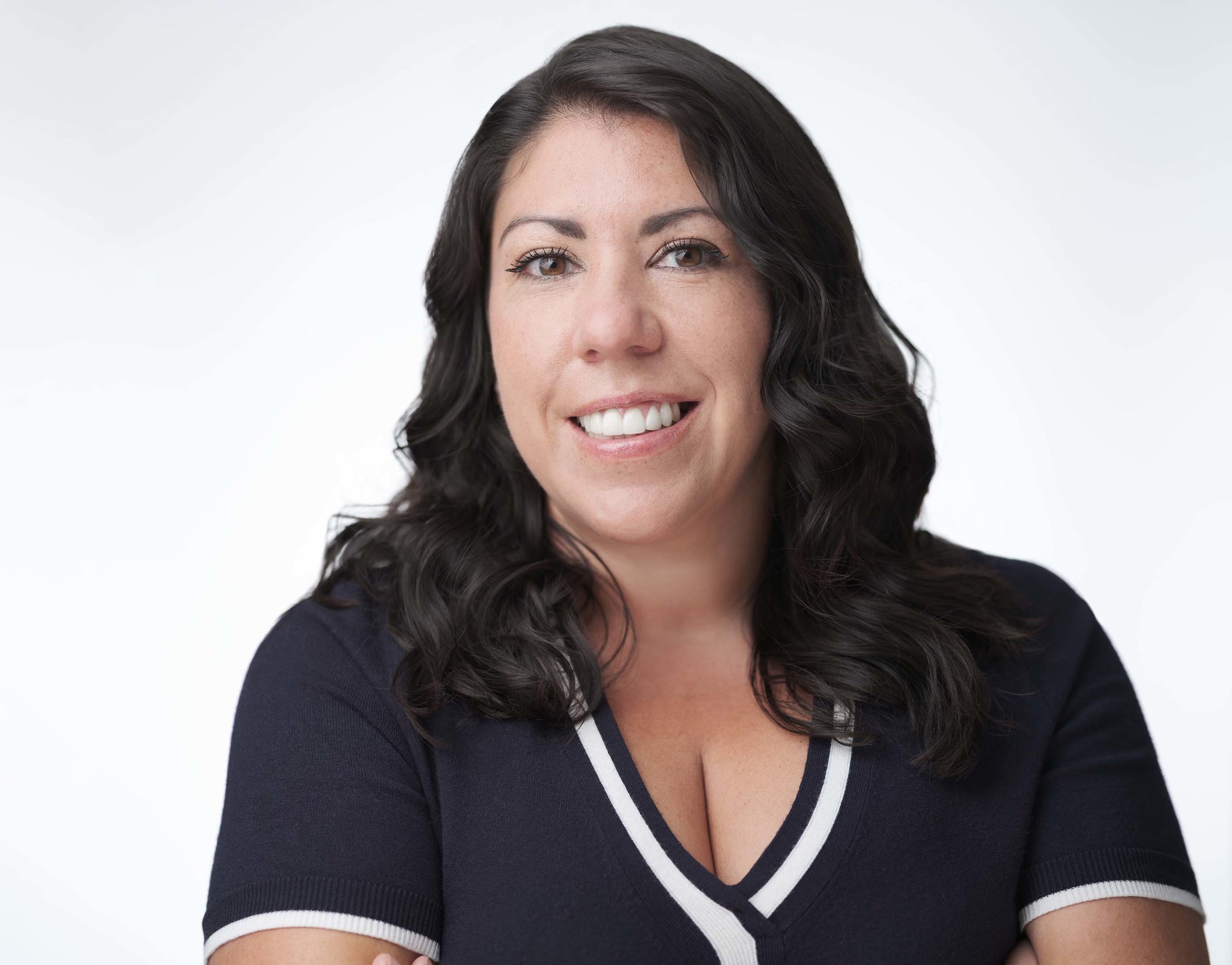 Woman smiling with dark, wavy hair, wearing a black shirt with white trim. She poses against a simple, light background.