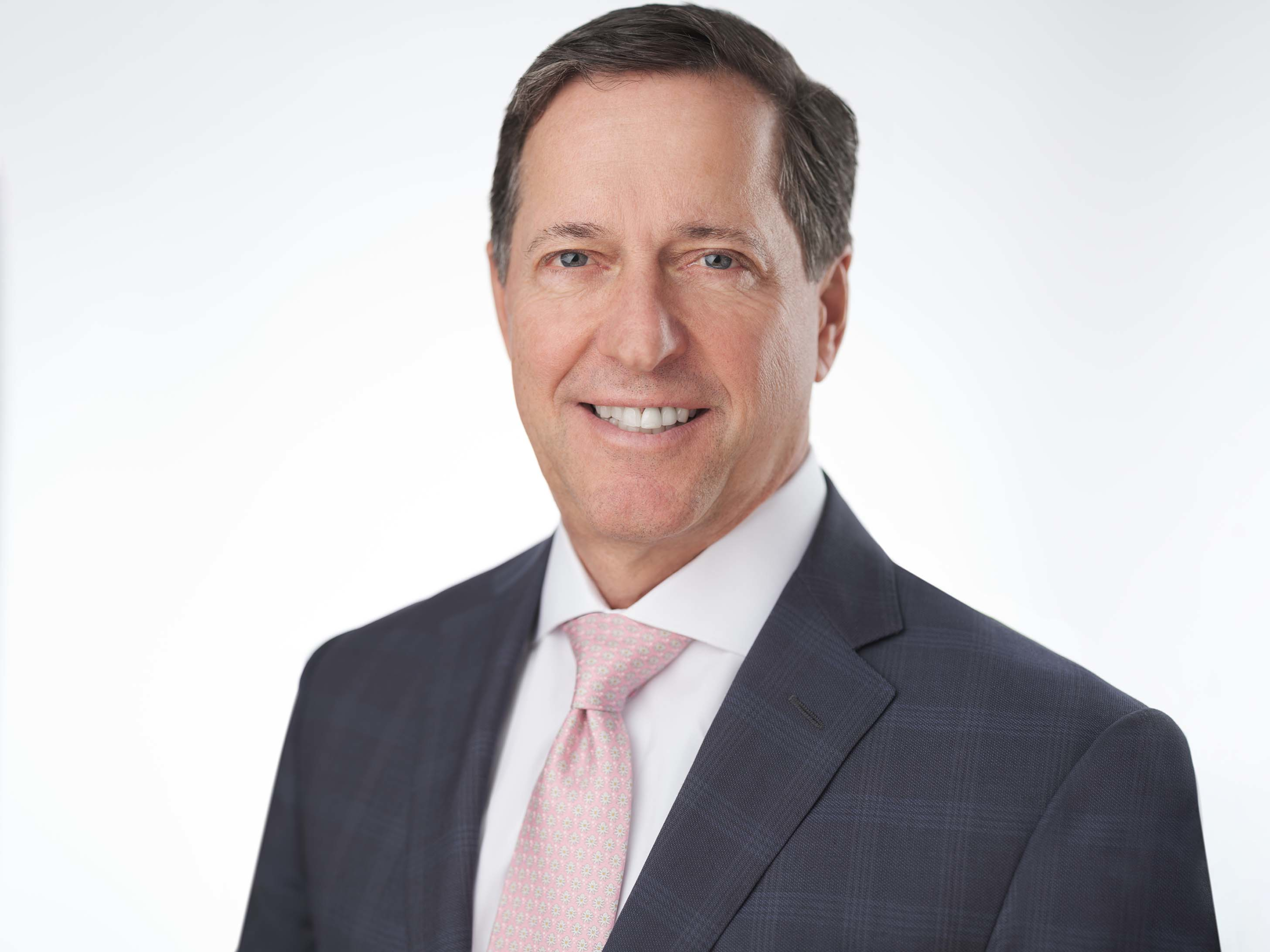 A man in a dark suit and light pink tie smiles at the camera against a plain white background.