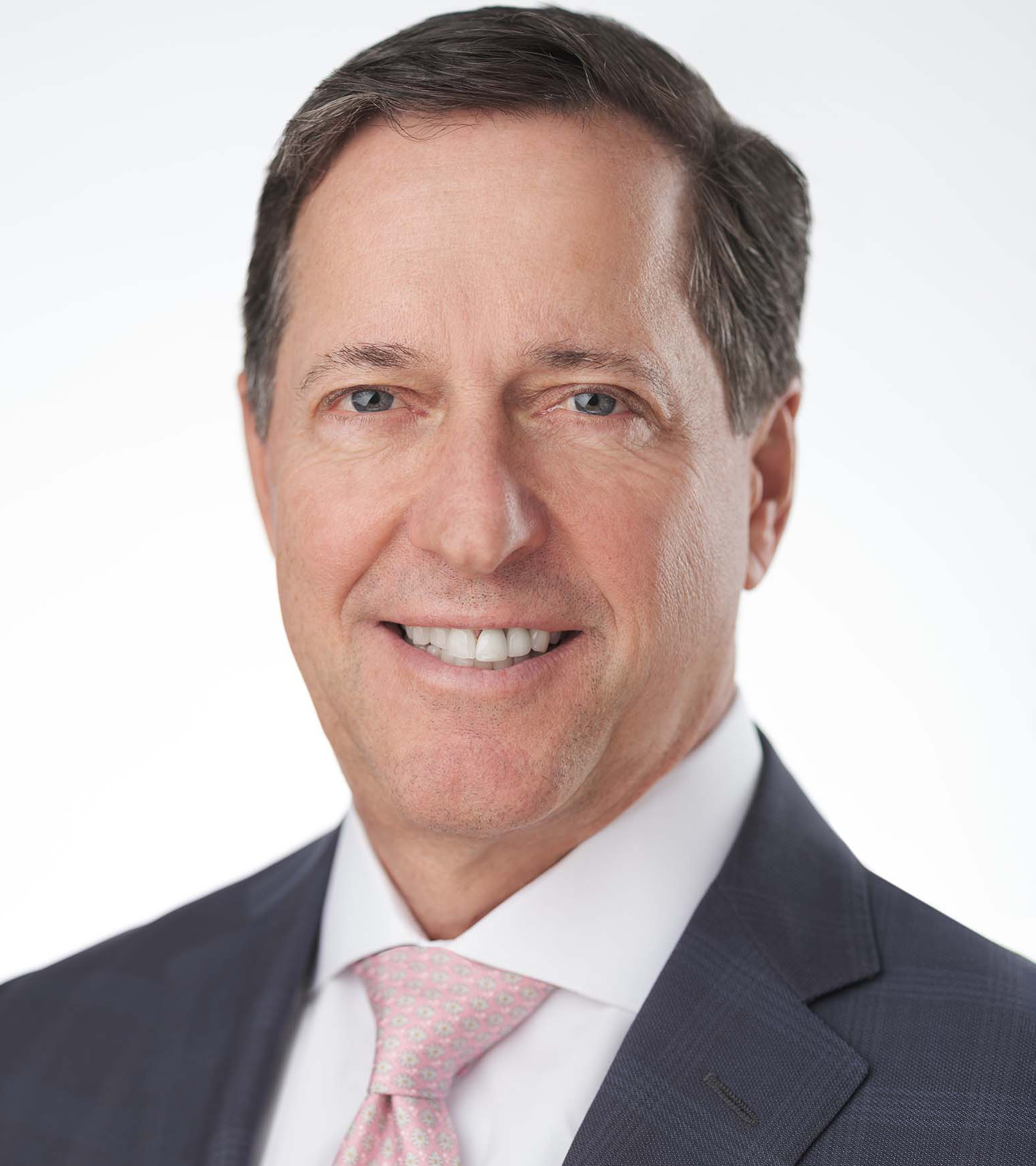 A man in a suit and pink tie smiles against a plain white background. He has short, dark hair and is wearing a white shirt.