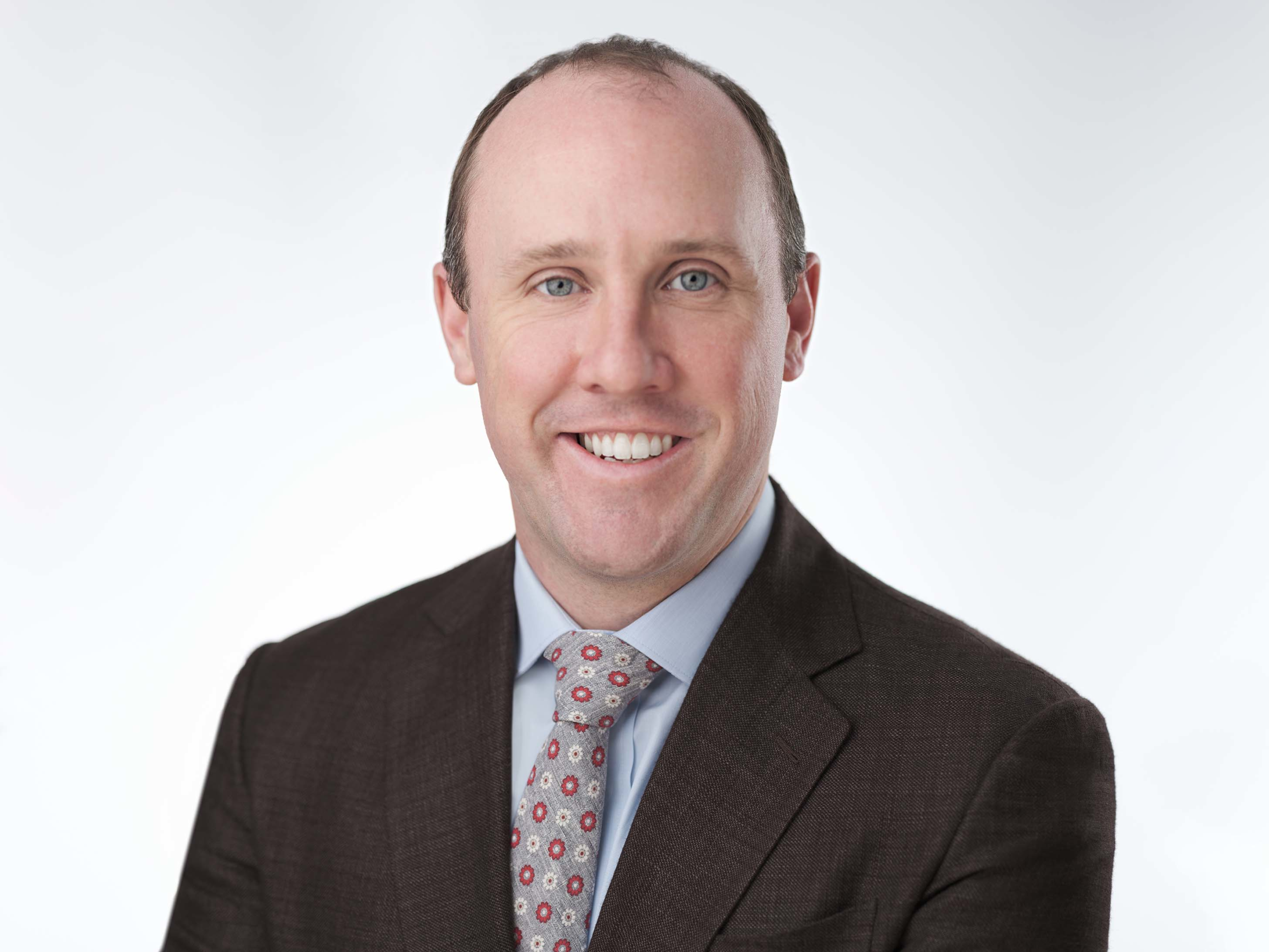 A person in a dark suit jacket, light blue dress shirt, and a patterned tie is smiling. The background is plain white, giving a professional headshot appearance.