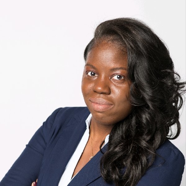 A person with long dark hair, wearing a navy blue blazer and a light-colored shirt, posing confidently with arms crossed against a plain white background.