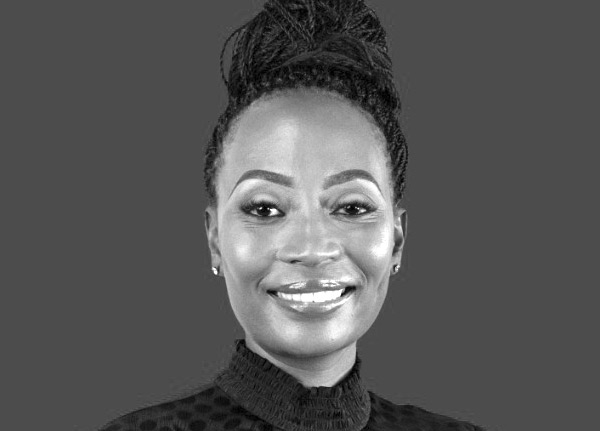 Black and white portrait of a smiling person with braided hair styled in an updo, wearing a high-necked, textured top, set against a plain dark background.