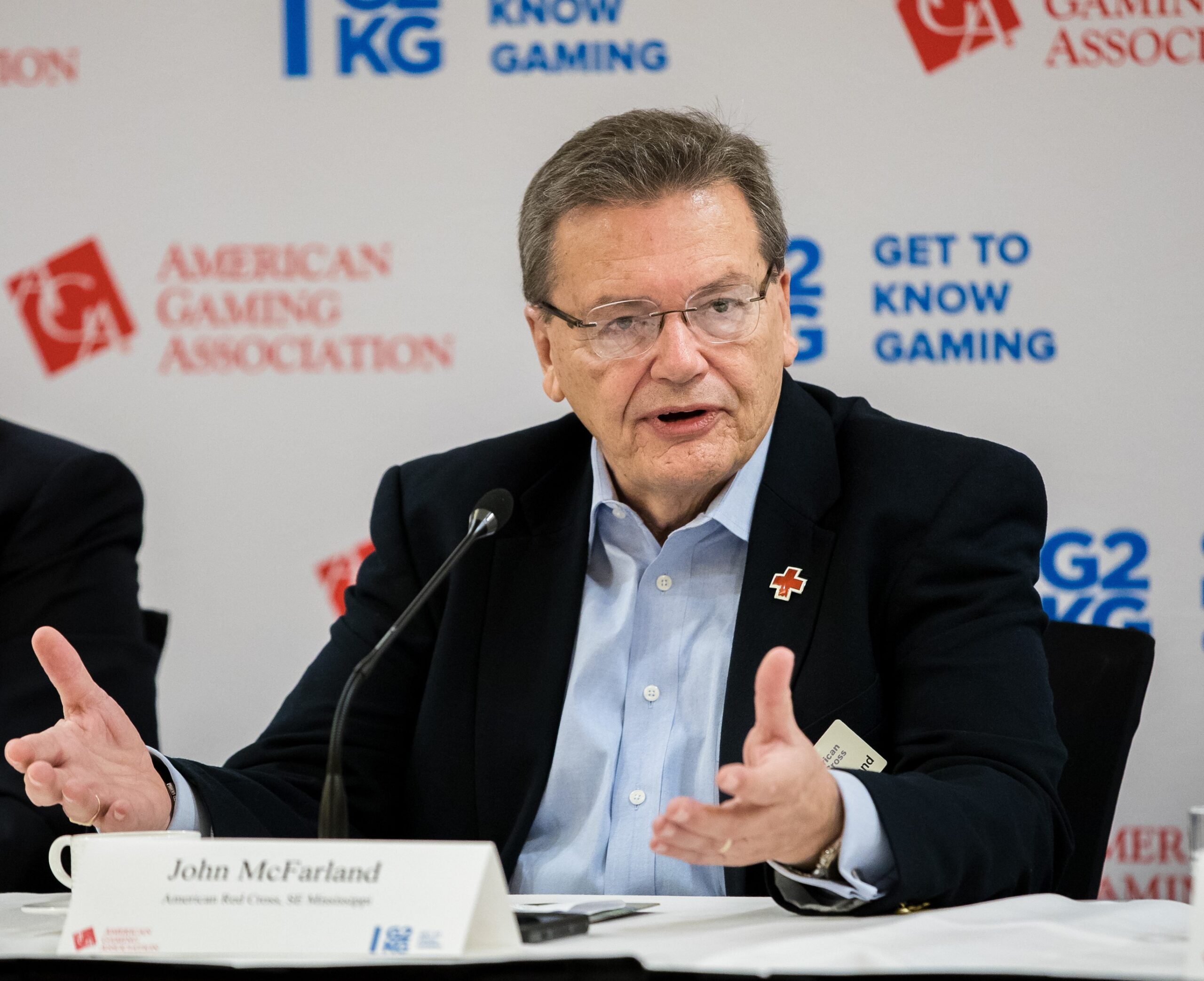 A man in a suit speaking at a microphone during a conference. He gestures with his hands. The background features logos of the American Gaming Association and "Get to Know Gaming." A name card in front of him reads "John McFarland.