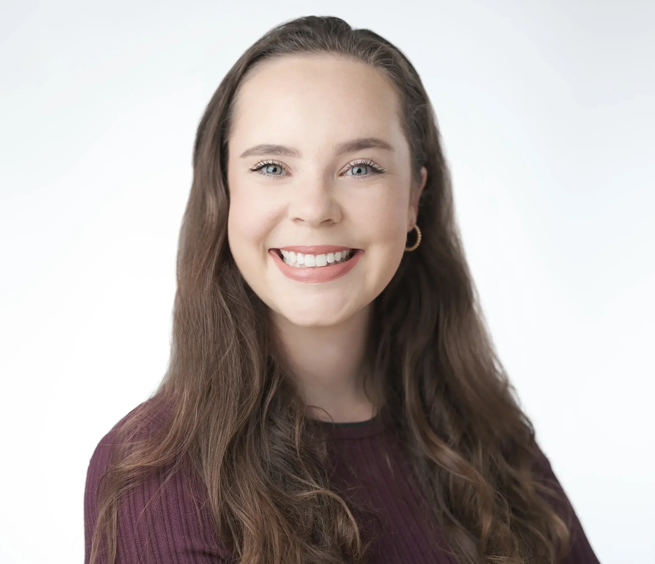 Smiling woman with long brown hair wearing a dark purple sweater and gold hoop earrings, posing against a light gray background.