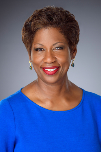 A woman wearing a bright blue top and green earrings smiles against a gray background. Her short hair is styled neatly.