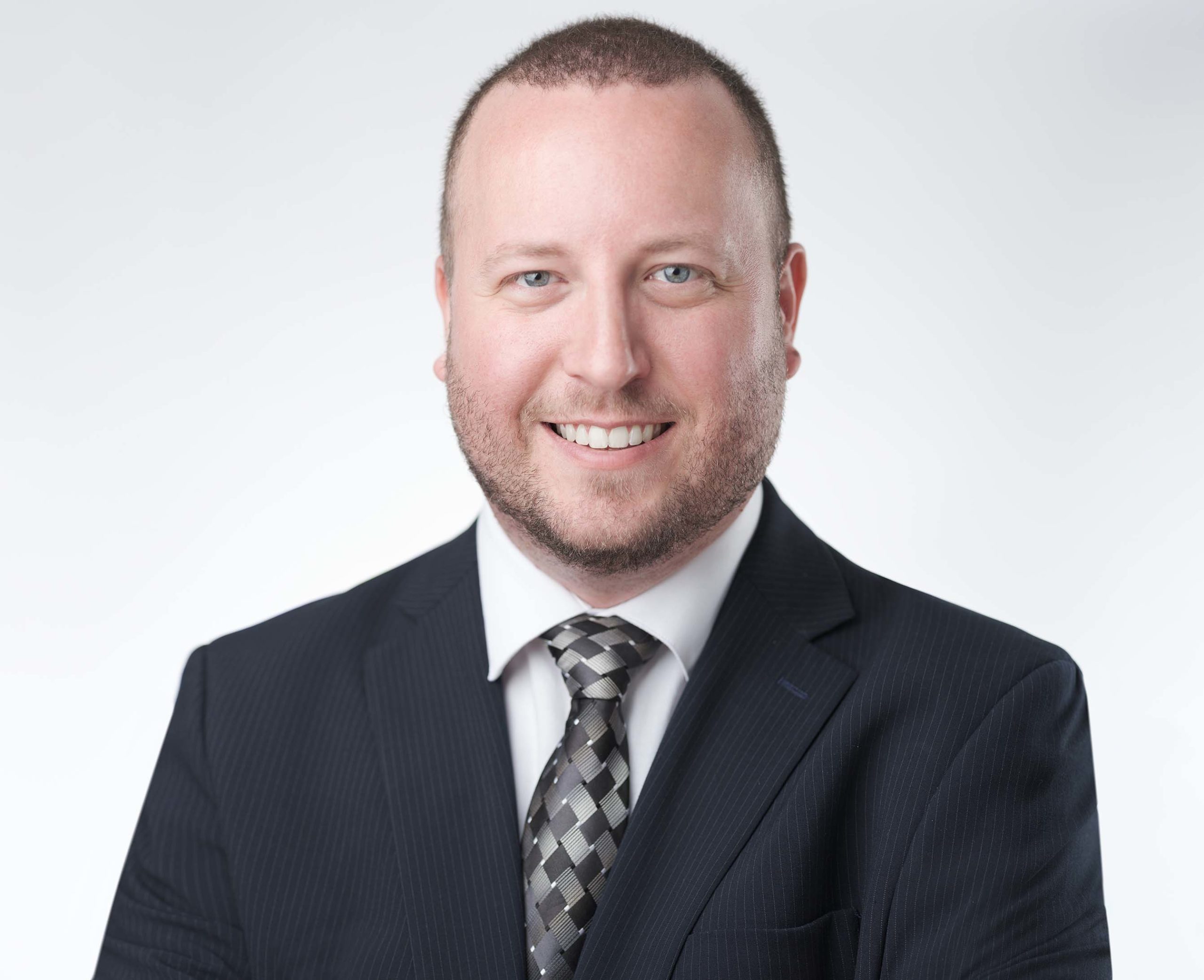A man with short hair and a beard smiles while wearing a dark suit, white shirt, and patterned tie. The background is a plain, light color.