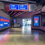 A stadium corridor with directional signs for sections 117-119 and suite 122. Digital displays feature health and gaming plan advertisements. A Dunkin' Donuts store is on the right, and an entrance with natural light is ahead.
