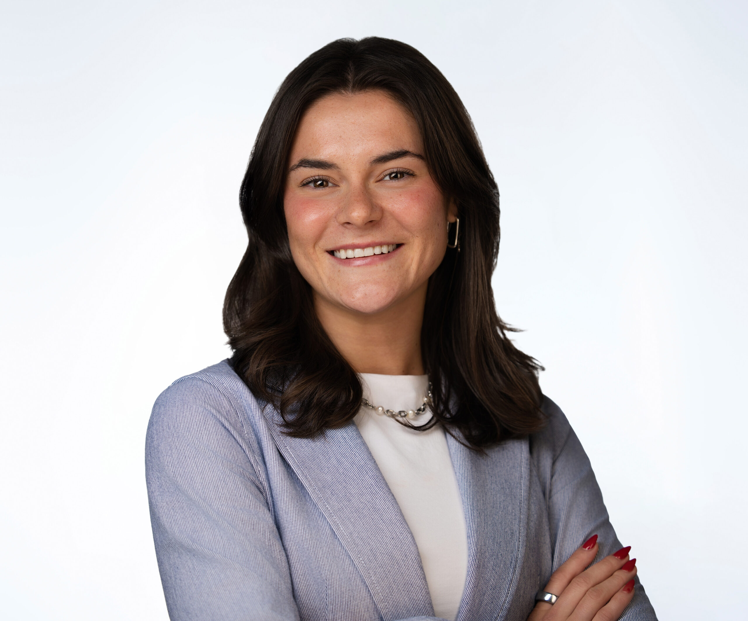 Smiling woman with dark shoulder-length hair, wearing a light blue blazer, white top, necklace, and red nail polish, poses with arms crossed against a plain white background.