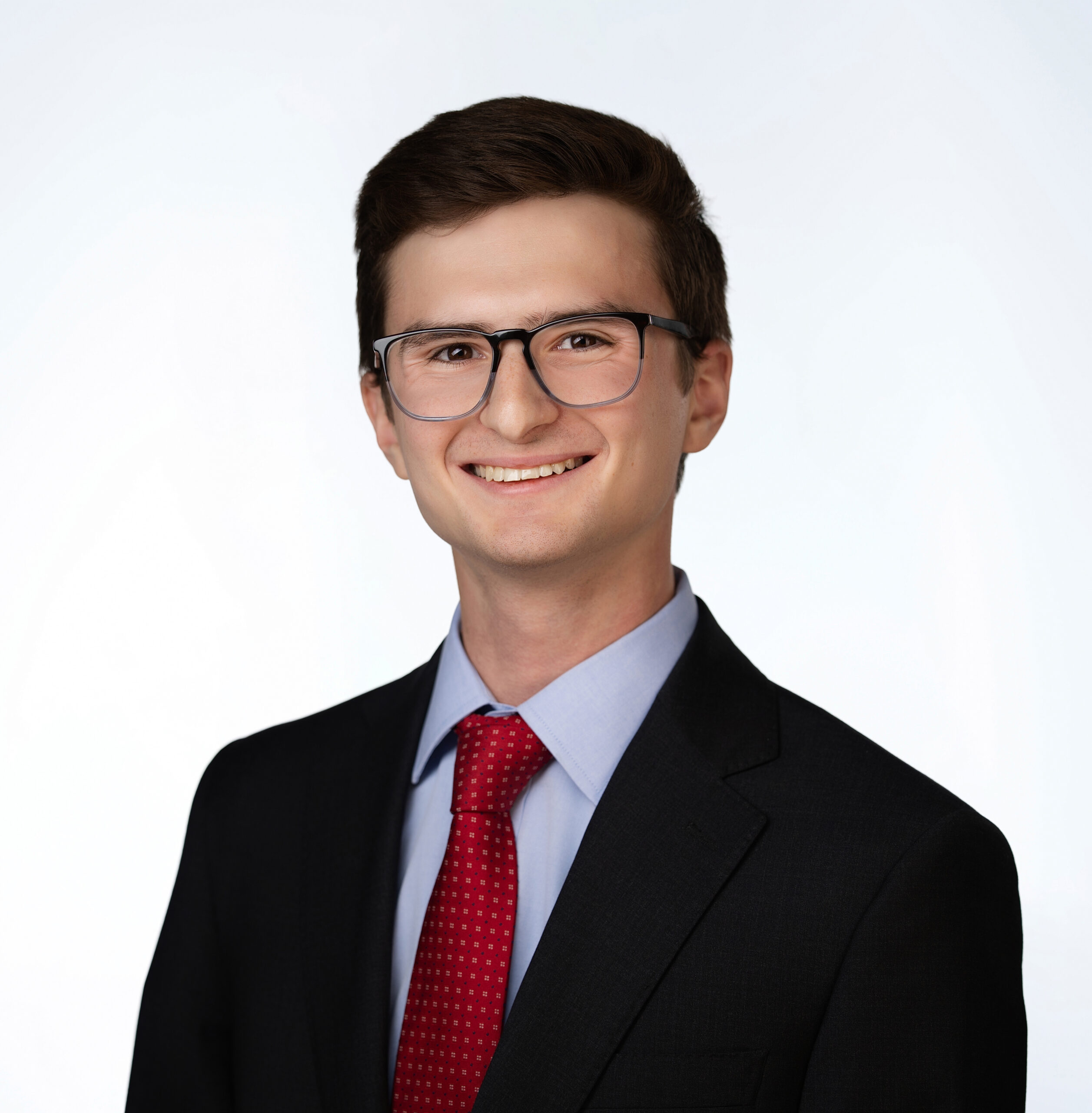 A young man with short brown hair and glasses is smiling, wearing a black suit, light blue shirt, and red tie, posed against a plain white background.