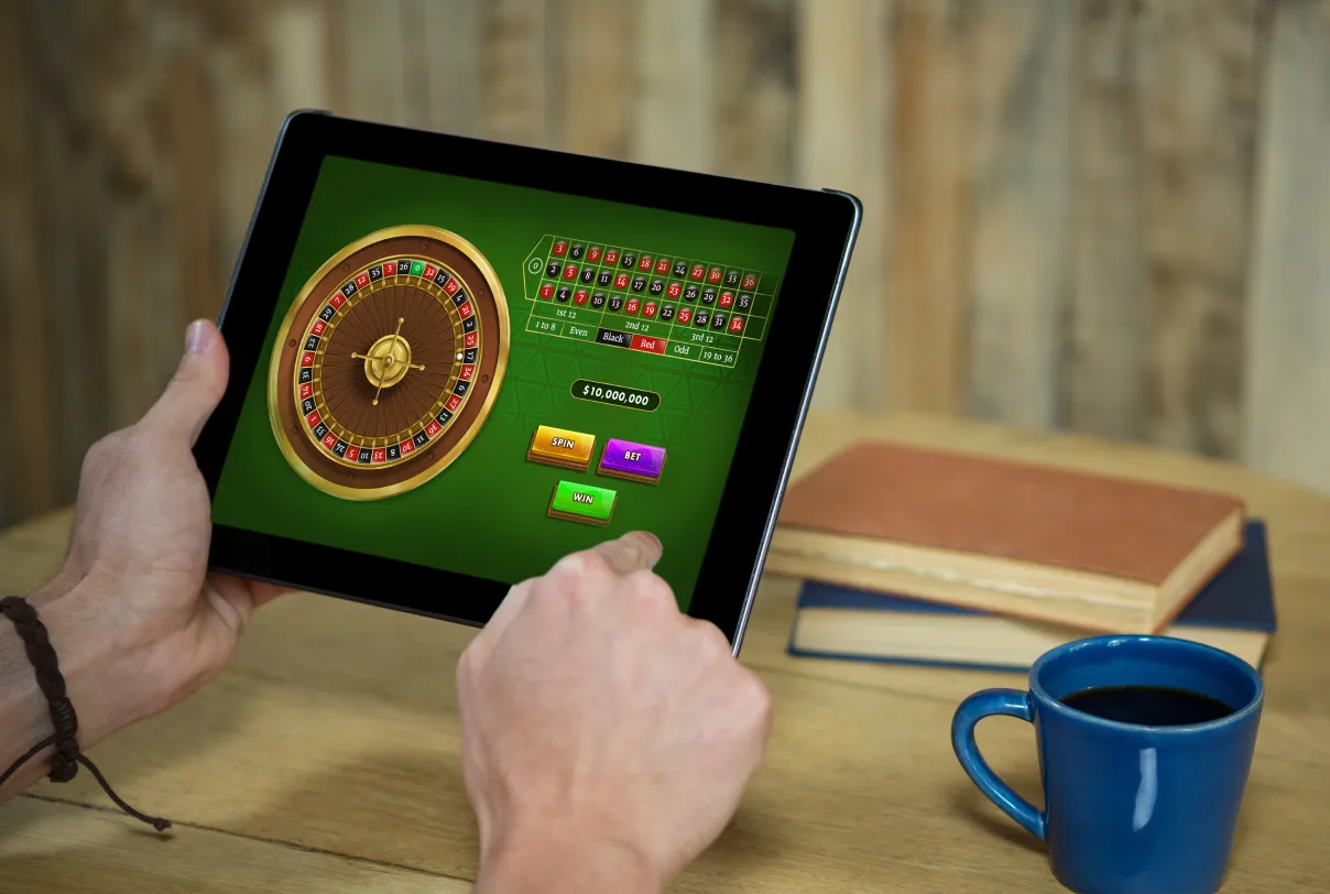 A person is playing a virtual roulette game on a tablet at a wooden table with a blue coffee cup, a closed notebook, and a book in the background.