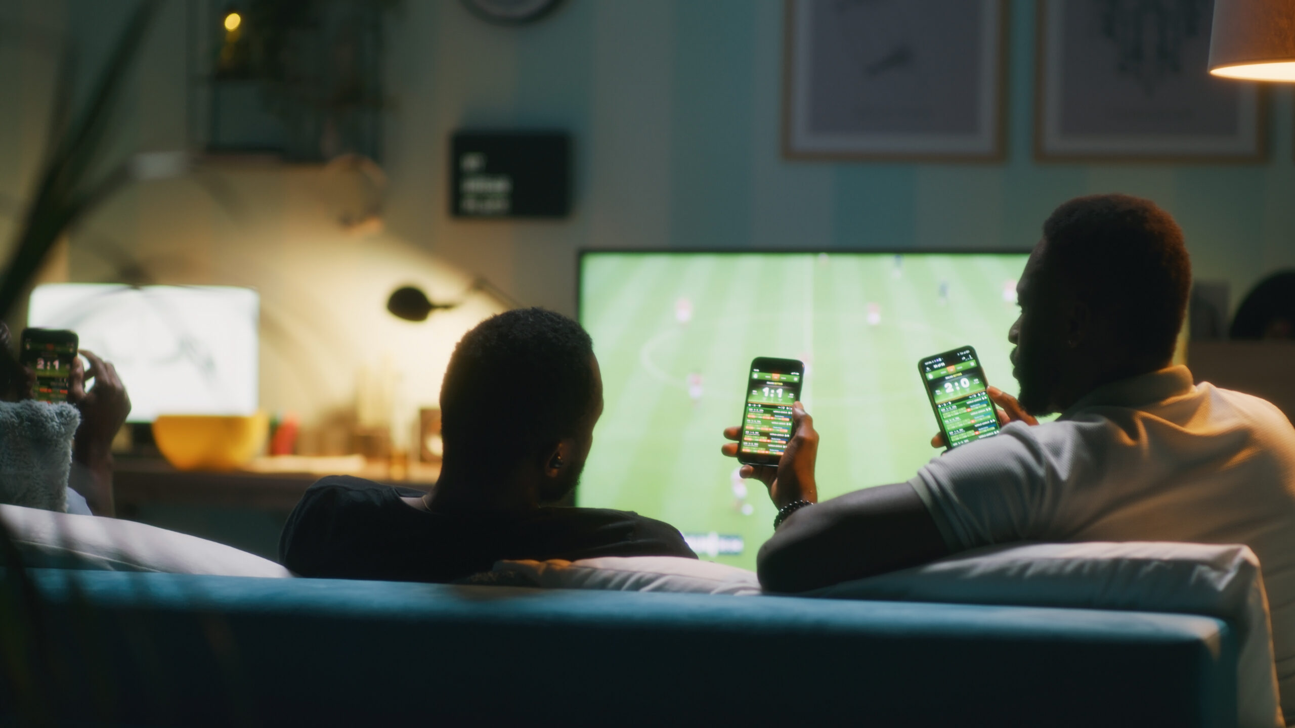 Two people sitting on a couch watching a soccer match on TV, both holding smartphones displaying illegal gambling apps, in a cozy, dimly lit living room.
